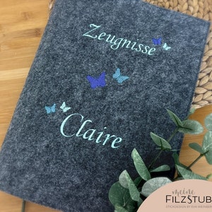 May include: Gray felt folder with the text "Zeugnisse" and "Claire" embroidered in white. The folder is decorated with blue and turquoise butterflies.