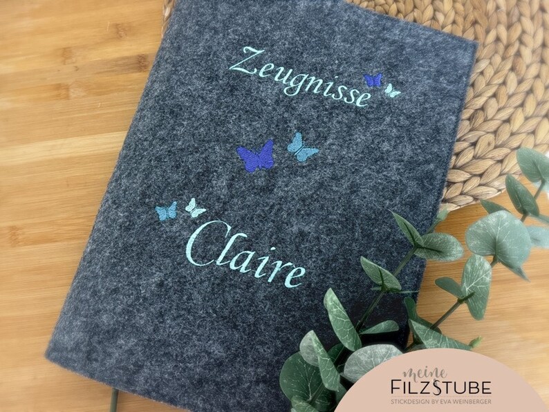 May include: Gray felt folder with the text "Zeugnisse" and "Claire" embroidered in white. The folder is decorated with blue and turquoise butterflies.