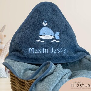 May include: A blue hooded towel with a whale and waves embroidered on it. The towel is personalized with the name "Maxim Jasper".