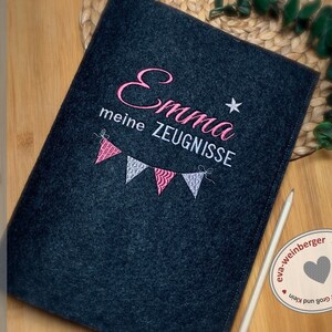 Pu&ograve; includere: Una cartella in feltro grigio con il testo "Emma meine ZEUGNISSE" ricamato in rosa e bianco. Una fila di bandierine rosa e bianche &egrave; ricamata sotto il testo. La cartella &egrave; su una superficie di legno.