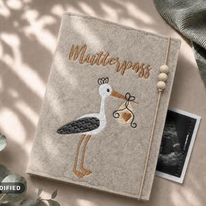 May include: A beige felt cover for a Mutterpass featuring an embroidered stork carrying a bundle with a heart. The cover includes a vertical elastic band with three wooden beads and displays the text Mutterpass in brown cursive embroidery.
