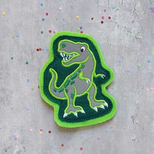 Könnte beinhalten: Grüner Filz-Dinosaurier-Patch mit einem grauen gestickten Dinosaurier. Der Dinosaurier hat einen grünen Umriss und einen weißen Mund.