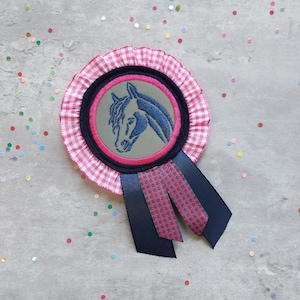 Könnte beinhalten: Eine rosa-weiße Gingham-Rosette mit einem blauen und grauen gestickten Pferdekopf. Die Rosette hat ein dunkelblaues Band.
