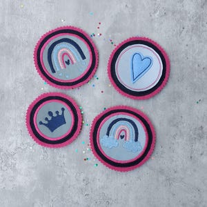 Könnte beinhalten: Vier runde, dekorative Aufnäher mit gestickten Designs. Jeder Aufnäher hat einen rosa, gewellten Rand, einen schwarz-weißen Rand und eine graue Mitte. Zu den Designs gehören ein Regenbogen, ein Herz, eine Krone und ein Regenbogen mit Wolken.