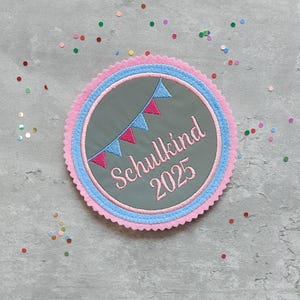 Könnte beinhalten: Runder, gestickter Aufnäher mit dem Text "Schulkind 2025". Der Aufnäher hat einen grauen Mittelteil, einen rosa und blauen Rand und eine Girlande aus rosa und blauen Fähnchen. Der Hintergrund ist grau mit bunten Konfetti.