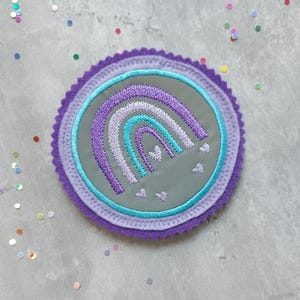 Könnte beinhalten: Ein gestickter Regenbogen-Patch in Lila und Blau mit weißen Herzen auf grauem Hintergrund. Der Patch ist von einem lilafarbenen Rand mit gewelltem Rand umgeben.