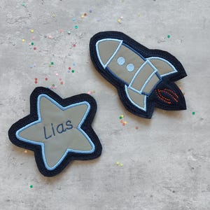 Könnte beinhalten: Zwei Filzpatches, einer ein blauer Stern mit dem Namen "Liam" bestickt, der andere eine blaue und graue Rakete mit einer orangefarbenen Flamme.