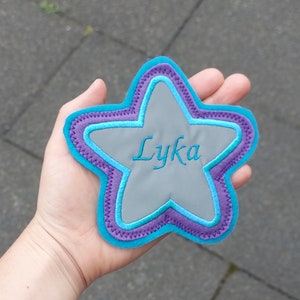 Könnte beinhalten: Ein blauer und violetter sternförmiger Patch mit dem Namen "Lyka", der in Weiß gestickt ist.