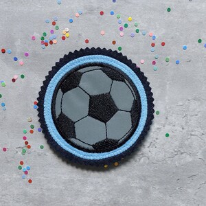 Könnte beinhalten: Ein kreisförmiger Aufnäher mit einem Fußball-Design. Der Ball ist grau und schwarz, mit einem hellblauen Ring und einem dunkelblauen, gewellten Rand. Bunte Konfetti sind um den Aufnäher auf einer grauen Oberfläche verstreut.