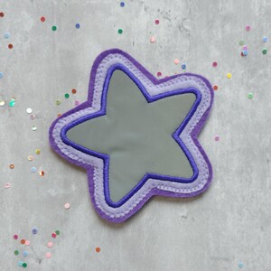 Könnte beinhalten: Ein sternförmiger Patch in Lila mit einem grauen Zentrum und einem doppelten lilafarbenen Umriss. Der Patch befindet sich auf einem grauen Hintergrund mit buntem Konfetti.