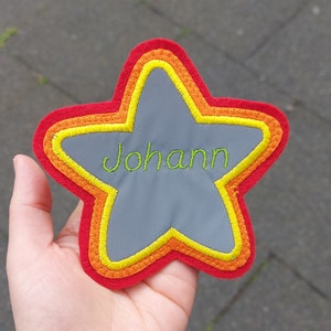 Könnte beinhalten: Ein roter sternförmiger Patch mit gelbem und orangefarbenem Besatz. Der Stern ist aus reflektierendem Material und trägt den Namen "Johann" in grüner Stickerei.