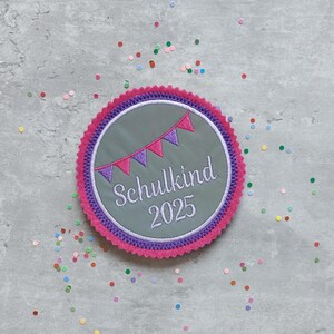 Könnte beinhalten: Ein runder, grauer Filz-Patch mit rosa und lila Besatz. Der Patch hat eine Girlande aus rosa und lila Flaggen und den Text "Schulkind 2025" in weißer Stickerei.