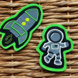 Könnte beinhalten: Zwei Filzpatches mit grünen Umrissen. Ein Patch zeigt ein graues Raumschiff mit gelben Details. Der andere Patch zeigt einen grauen Astronauten mit einem schwarzen Helm und einem grünen Visier.