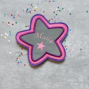 Könnte beinhalten: Ein sternförmiger Aufnäher mit grauem Zentrum, bestickt mit dem Namen "Maila" in Schreibschrift und einem kleinen Stern. Der Aufnäher hat einen rosa-lila-farbenen Rand. Buntes Konfetti ist um den Aufnäher verstreut.