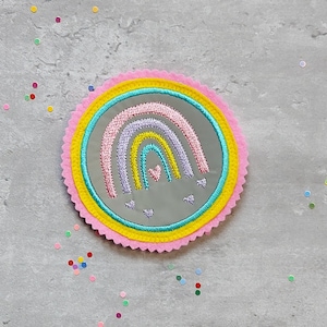 Könnte beinhalten: Ein rosa, gelber und blauer Regenbogen mit Herzen, gestickt auf einem silbernen reflektierenden Stoffpatch. Der Patch ist von einem rosa, gelben und blauen Besatz umgeben.