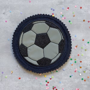 Könnte beinhalten: Ein gestickter Fußball-Patch in Schwarz und Grau mit einem dunkelblauen Rand. Der Patch befindet sich auf einem grauen Hintergrund mit buntem Konfetti.