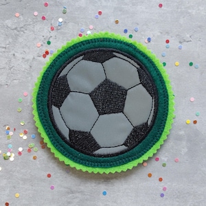 Könnte beinhalten: Grün-schwarze Filzapplikation eines Fußballs mit weißem Hintergrund. Die Applikation hat einen gewellten Rand.