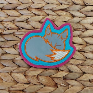 Könnte beinhalten: Ein gestickter Aufnäher mit einem schlafenden Fuchs, mit einem rosa und blauen Rand.
