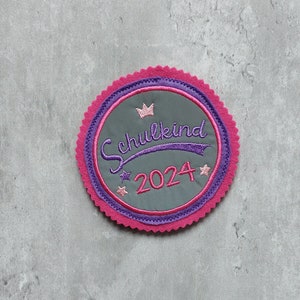 Könnte beinhalten: Ein rosa-lila gestickter Aufnäher mit einer Krone und dem Text "Schulkind 2024" in lila.