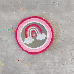 Könnte beinhalten: Ein rosa-weißer Regenbogen mit Wolken, der auf einen grauen Filzkreis mit rosa Rand gestickt ist. Der Kreis ist von buntem Konfetti umgeben.