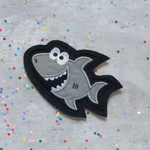 Könnte beinhalten: Ein grauer Filz-Hai-Patch mit weißen Augen und einem schwarzen Umriss. Der Hai lächelt mit offenem Maul und hat eine Flosse auf dem Rücken und am Schwanz.
