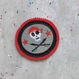 Könnte beinhalten: Gestickter Piratenaufnäher mit einem Totenkopf mit rotem Bandana und Augenklappe, gekreuzten Schwertern und roten Sternakzenten. Der Aufnäher hat einen grauen Hintergrund, einen schwarzen Rand und einen roten, gewellten Rand. Ein lustiges Accessoire für Kleidung oder Taschen.