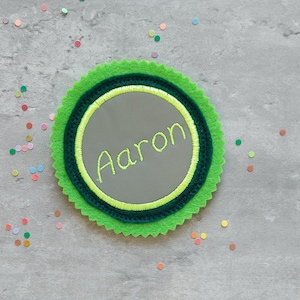 Könnte beinhalten: Ein grüner Filzkreis mit einem gewellten Rand und einem kleineren Kreis im Inneren. Der kleinere Kreis ist grau und hat den Namen "Aaron" in gelber Farbe gestickt.