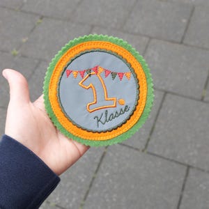 Könnte beinhalten: Ein runder, grauer Filz-Patch mit grünem und orangefarbenem Rand. Der Patch hat eine große gestickte Zahl "1" mit einer Flaggenkette darüber. Der Text "Klasse" ist unter der Zahl gestickt.