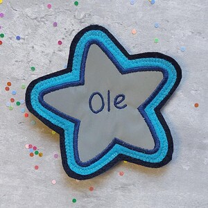 Könnte beinhalten: Ein blauer und grauer sternförmiger Patch mit dem Namen "Ole", der in weißer Farbe gestickt ist.