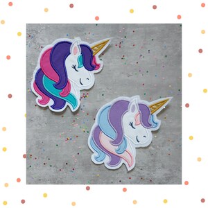 Könnte beinhalten: Zwei gestickte Bügelbilder mit Einhorn-Motiven. Die Einhörner sind weiß mit lila, rosa und blauen Mähnen und Schwänzen. Die Einhörner haben goldene Hörner.