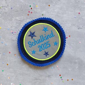 Könnte beinhalten: Ein blauer und schwarzer runder Patch mit einem grauen Zentrum. Das Zentrum trägt den Text "Schulkind 2025" in blau mit fünf blauen Sternen um den Text. Der Patch hat einen gelben Rand.