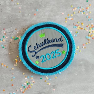 Könnte beinhalten: Ein runder, gestickter Aufnäher mit dem Text "Schulkind 2025" in marineblauer Schrift. Der Aufnäher hat eine graue Mitte, einen marineblauen Ring und einen hellblauen, gewellten Rand. Grüne Sterne und eine Krone akzentuieren das Design.
