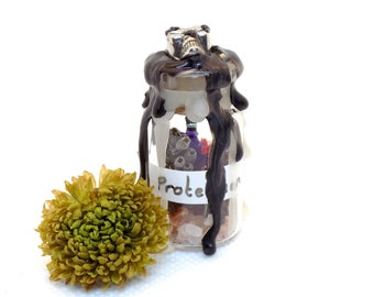 May include: A small glass bottle filled with various crystals and herbs, sealed with black wax and a silver skull charm. The bottle is labelled "Protection" and is sitting on a white surface next to a dried green flower.