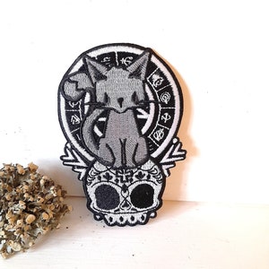 May include: Black and white embroidered patch featuring a cat sitting on a skull with a circle of symbols around the cat.