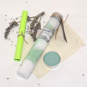Pode incluir: Um tubo de vidro cheio de sais de banho brancos e verdes, uma pequena vela turquesa, um papel verde enrolado e uma pequena bolsa de tecido branco com um cordão marrom.