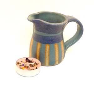 May include: A blue and brown striped ceramic jug with a handle. A small pink candle with dried flowers and herbs is in the foreground.