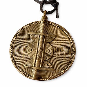 May include: A round, gold-colored pendant with a central design and spiral detailing. The pendant is suspended from a dark brown leather cord. The design features a central vertical element with horizontal lines and a spiral pattern.