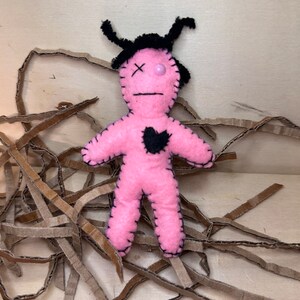 May include: Pink felt voodoo doll with black yarn hair, black eyes, and a black heart on its chest. The doll is sitting on brown shredded paper.