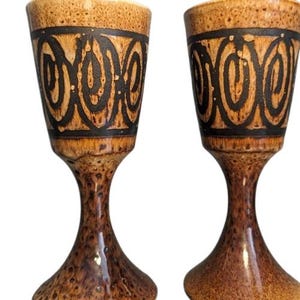 May include: Two brown ceramic shot glasses with black geometric designs. The glasses have a flared base and a rounded top.