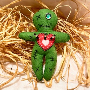 May include: Green felt voodoo doll with a red heart on its chest, surrounded by straw. The doll has black stitches for eyes and a green gem for one eye. The heart has white stitching and is pierced with black pins.