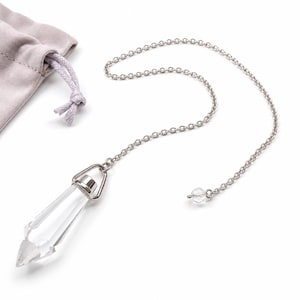 Pode incluir: Um pêndulo de cristal transparente com uma corrente prateada e uma pequena conta transparente. O pêndulo está ao lado de uma pequena bolsa de veludo roxo claro com fecho de cordão. O cristal é facetado e pontiagudo.