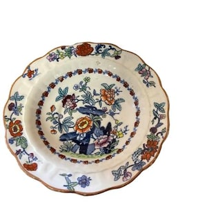 Puede incluir: Un plato de cerámica blanca con borde festoneado y borde marrón. El plato presenta un diseño floral con flores y hojas naranjas, azules, rosas y verdes. El plato es probablemente para fines decorativos o de servicio.