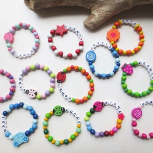 Peut inclure: Plusieurs bracelets de perles colorées avec des noms épelés en perles blanches. Chaque bracelet est orné d'un charm unique, comme des coccinelles, des fleurs et une voiture. Ces bracelets sont pour enfants.