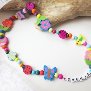 Könnte beinhalten: Eine bunte Holz Halskette mit Perlen, Blumen, Vögeln, Schmetterlingen, Marienkäfern und dem Namen "SVENJA" ausgeschrieben.