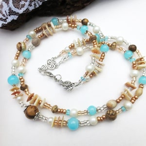 Op de afbeelding: Een gelaagde ketting met een mix van kralen in verschillende kleuren en texturen. De ketting bevat lichtblauwe, witte en bruine kralen, samen met kleine schelpachtige stukjes. De ketting is vastgemaakt met een zilveren sluiting.