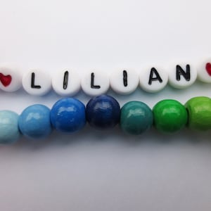 May include: A colorful wooden bead bracelet with the name "LILIAN" spelled out in white beads with black lettering. The bracelet has red heart beads on either side of the name.