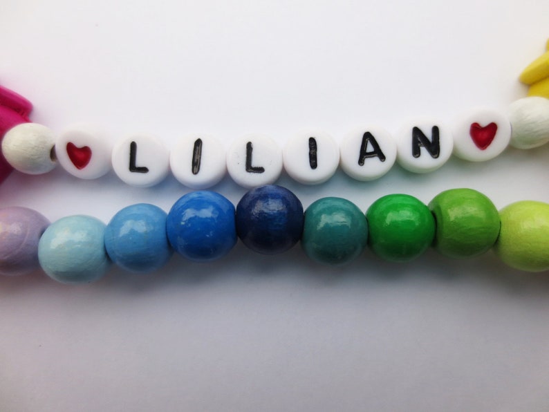 May include: A colorful wooden bead bracelet with the name "LILIAN" spelled out in white beads with black lettering. The bracelet has red heart beads on either side of the name.