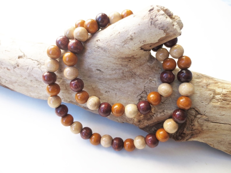 May include: A wooden bead necklace with brown, beige, and reddish-brown beads. The necklace is draped over a piece of driftwood.