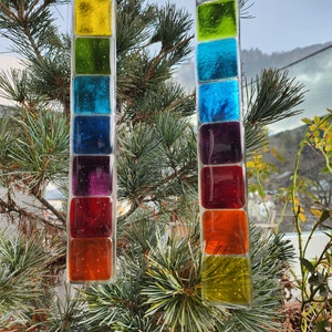 Op de afbeelding: Twee glazen zonvangers met een regenboog aan kleuren. Elke zonvanger heeft zeven vierkanten van glas in verschillende kleuren, waaronder geel, groen, blauw, paars, rood, oranje en geel. De zonvangers hangen aan een wit touwtje.