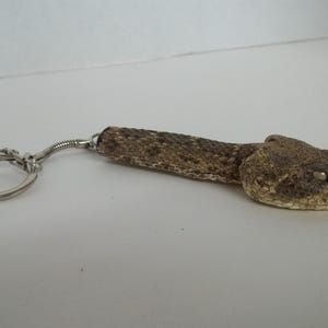 May include: A brown and tan rattlesnake keychain with a silver key ring. The snake is curled up with its head facing the camera.
